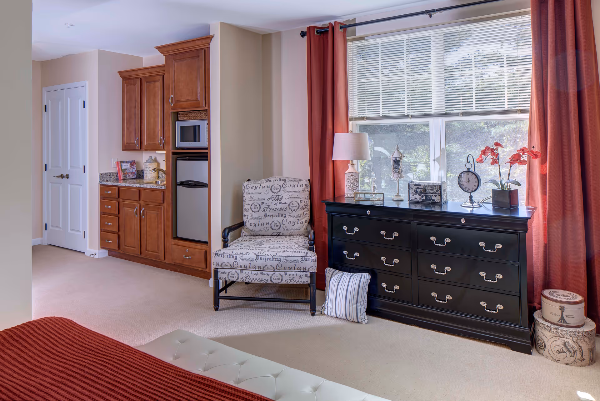 A cozy bedroom corner featuring a patterned armchair next to a black dresser with multiple drawers. The dresser holds decorative items including a table lamp, a clock, a jewelry stand, and a potted orchid. Large windows with white blinds and red curtains let in natural light. To the left, there is a built-in wooden cabinet with a microwave and mini fridge. The room has beige carpet and light-colored walls.