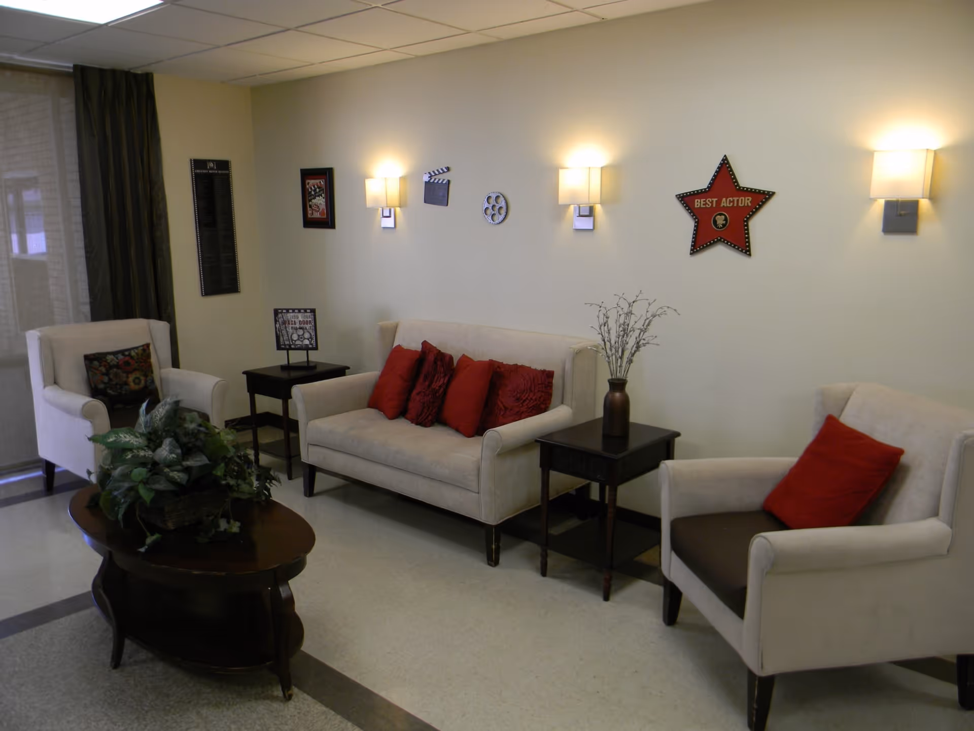 A cozy living room area with two beige armchairs and a beige loveseat, each adorned with red and patterned throw pillows. There are two dark wooden side tables, one with a decorative vase and the other with a framed picture. A dark wooden coffee table with a green plant centerpiece is in the foreground. The walls are decorated with movie-themed items including a red star plaque labeled 'Best Actor', a film reel, and a clapperboard. Soft wall-mounted lights illuminate the space.