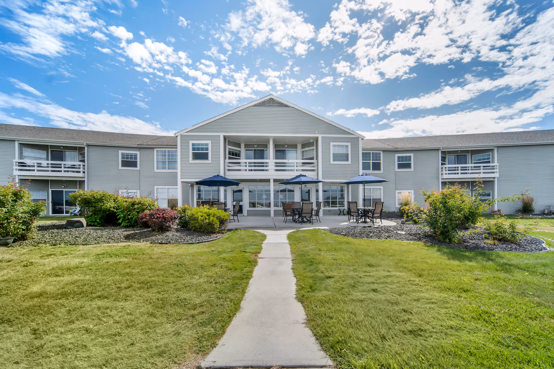 Exterior view of a two-story senior living facility building with balconies and a patio area featuring tables with umbrellas. The building is surrounded by well-maintained green lawns, shrubs, and a clear blue sky with scattered clouds.