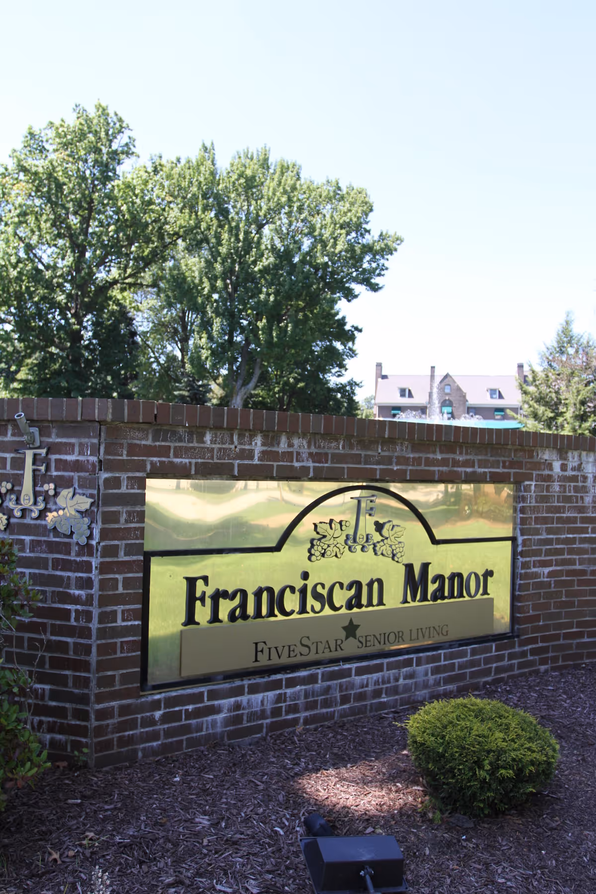 A brick sign for Franciscan Manor, a Five Star Senior Living facility, with trees and a building visible in the background under a clear sky.