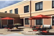 Outdoor patio area of a nursing and rehabilitation center with several round tables and chairs under red umbrellas. The building has beige walls and multiple windows.