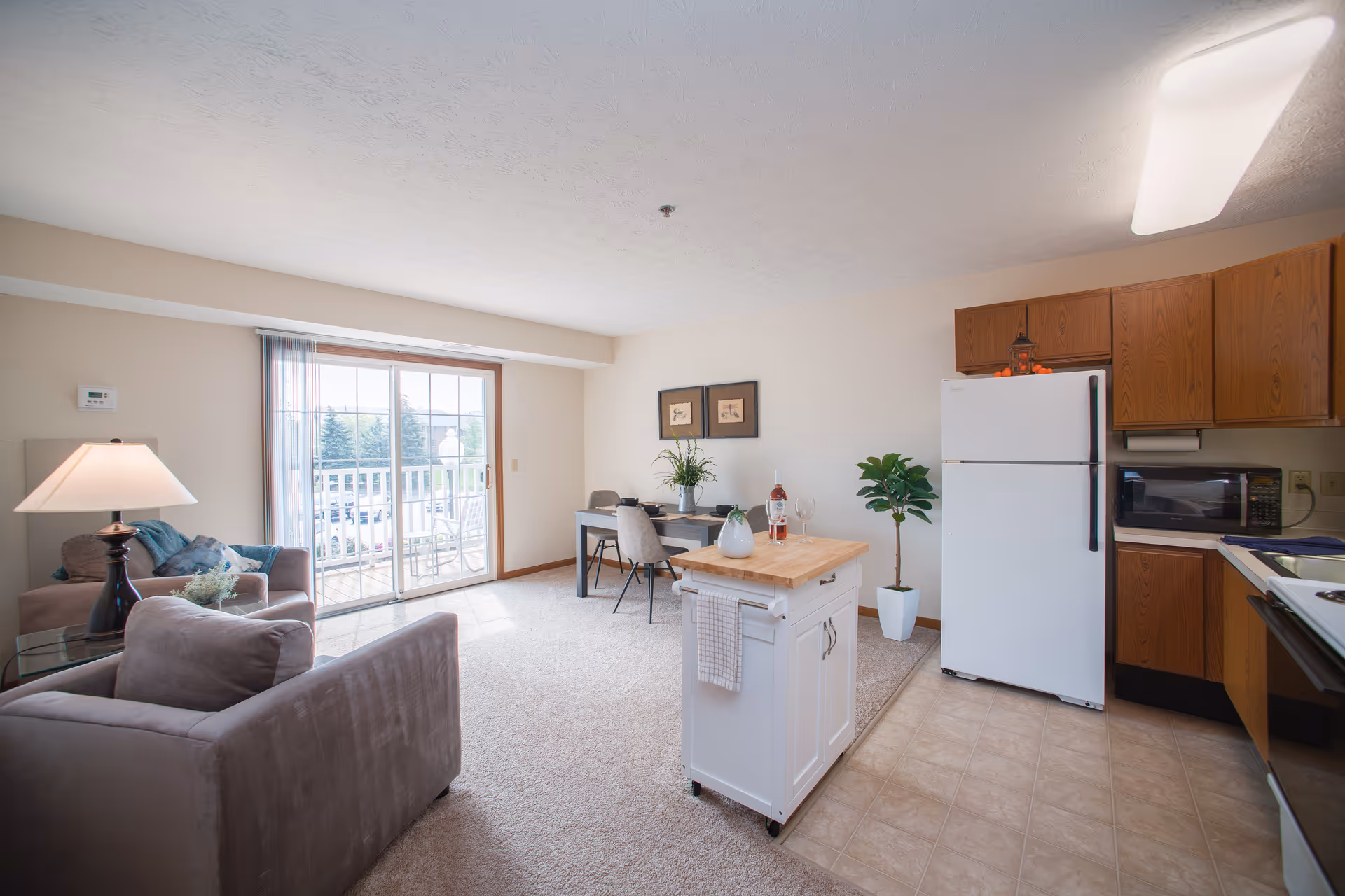 Open-plan apartment living area with sofas, a sliding glass door to a balcony, and a small kitchen with an island and white refrigerator.