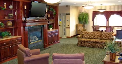 Cozy communal living room with upholstered sofas and chairs, a fireplace with a mounted TV and built-in shelving, and a dining area by the windows.