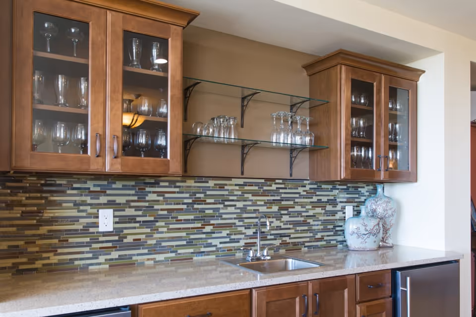 A kitchen area with wooden cabinets featuring glass doors displaying glassware, two glass shelves holding more glasses, a mosaic tile backsplash in shades of green, brown, and beige, a stainless steel sink with a faucet, and decorative ceramic vases on the countertop.