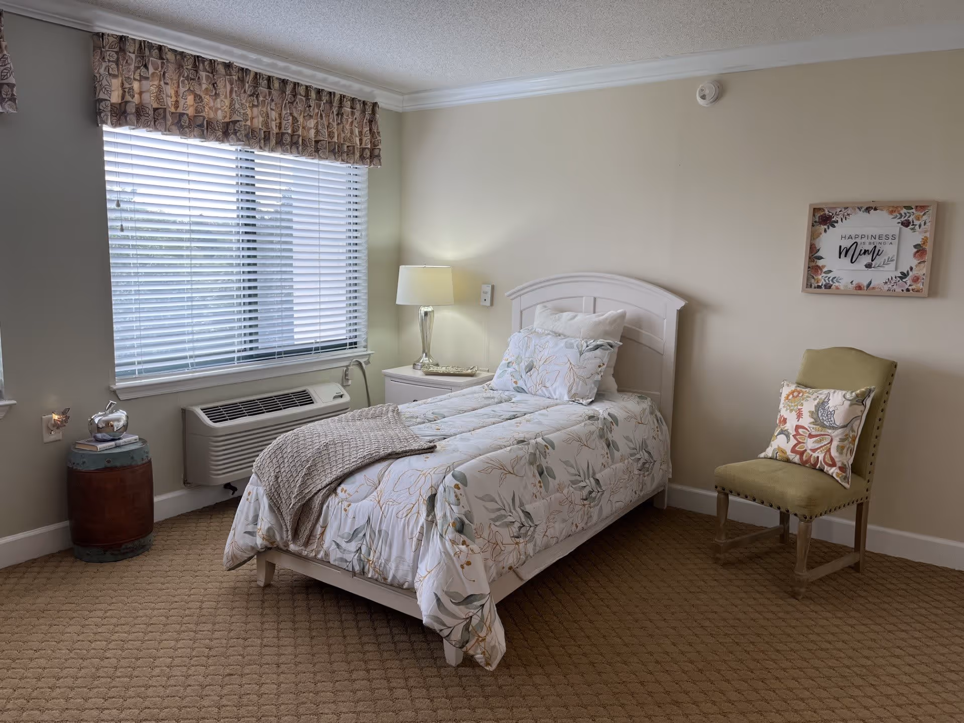 A cozy bedroom with a single bed covered in floral bedding and a knitted throw blanket. Next to the bed is a white nightstand with a lamp and decorative items. A window with blinds and a patterned valance lets in natural light. There is a green upholstered chair with a colorful pillow against the wall, which also features a framed floral print with the words 'Happiness is Homemade'. The room has beige carpet and light-colored walls.