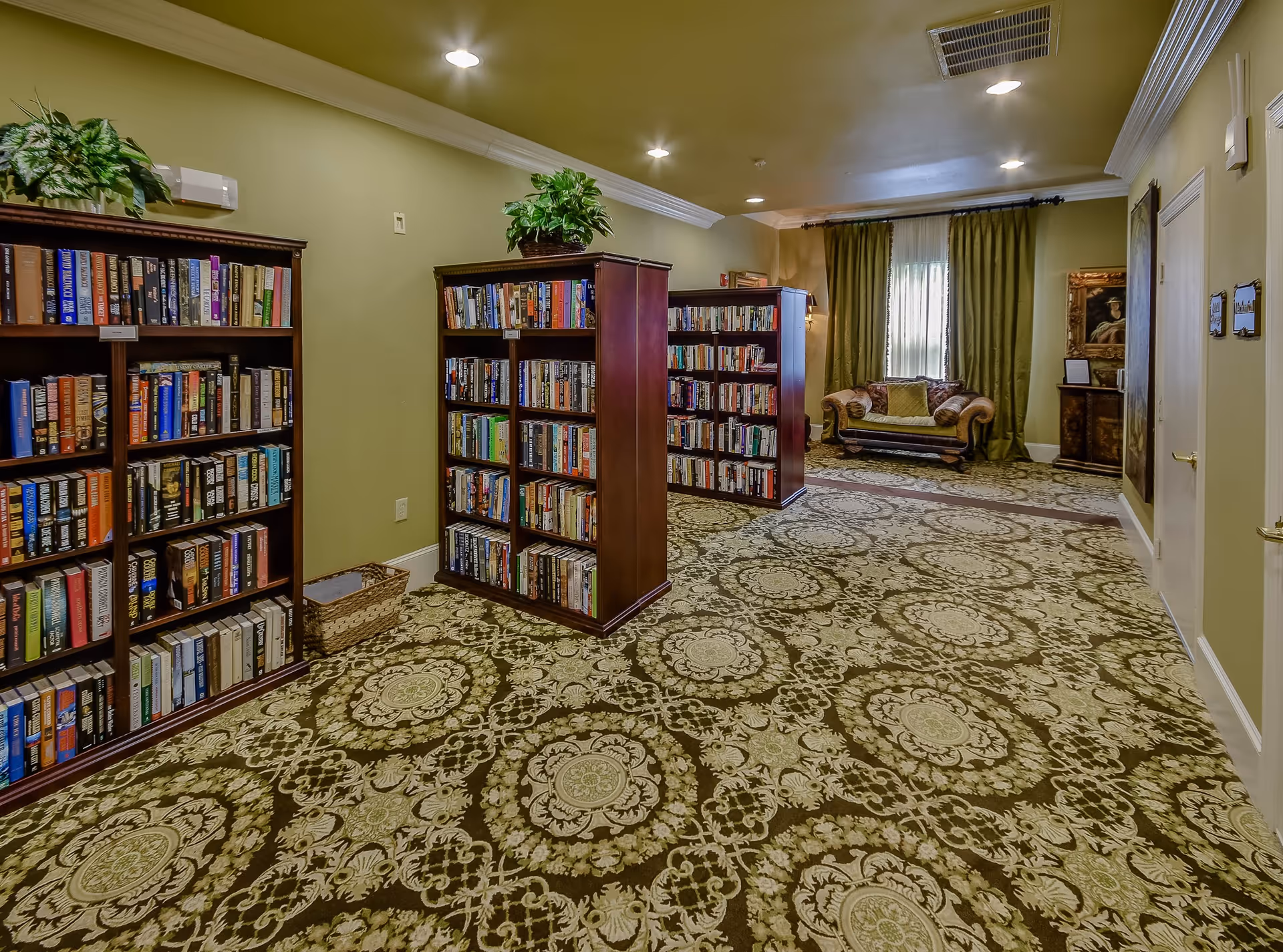 Interior view of a library or reading room with multiple wooden bookshelves filled with books, a patterned carpet, green walls, ceiling lights, and a small sofa with green curtains in the background.