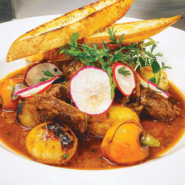 A plated dish featuring braised meat with grilled baby onions and small yellow vegetables in a rich brown sauce, garnished with thinly sliced radishes and fresh green herbs, accompanied by two slices of toasted baguette.