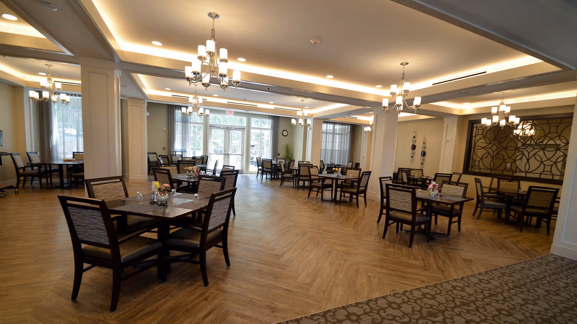 A spacious dining room with multiple square wooden tables and chairs arranged neatly. The room features wooden flooring, elegant chandeliers hanging from the ceiling, large windows with sheer curtains allowing natural light, and decorative wall elements.