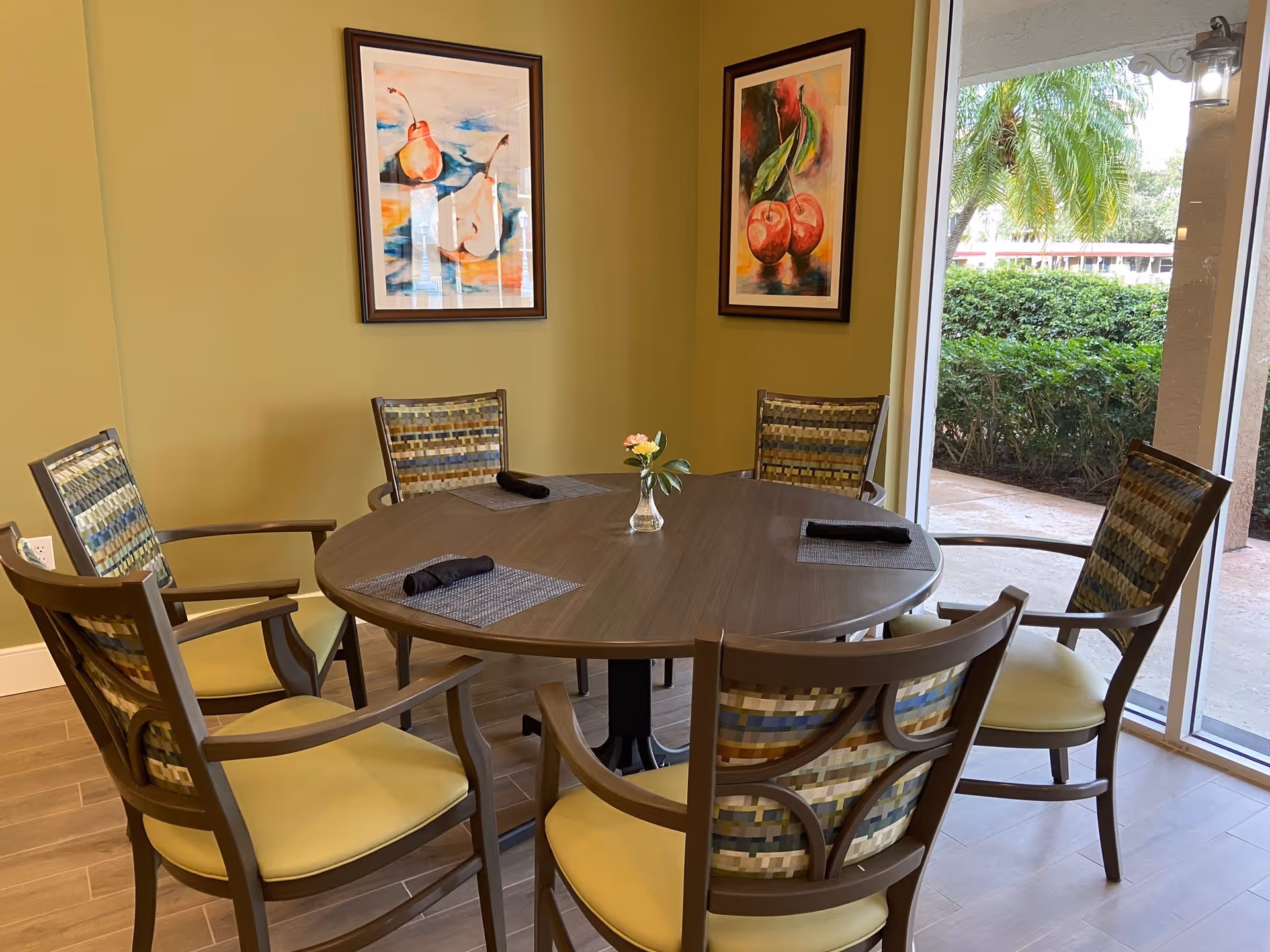 A round wooden dining table with six chairs around it, each chair having a patterned backrest and yellow seat cushion. The table has four placemats with black napkins and a small vase with flowers in the center. The room has light green walls with two framed paintings of fruit hanging on them. There is a large window showing greenery and palm trees outside.