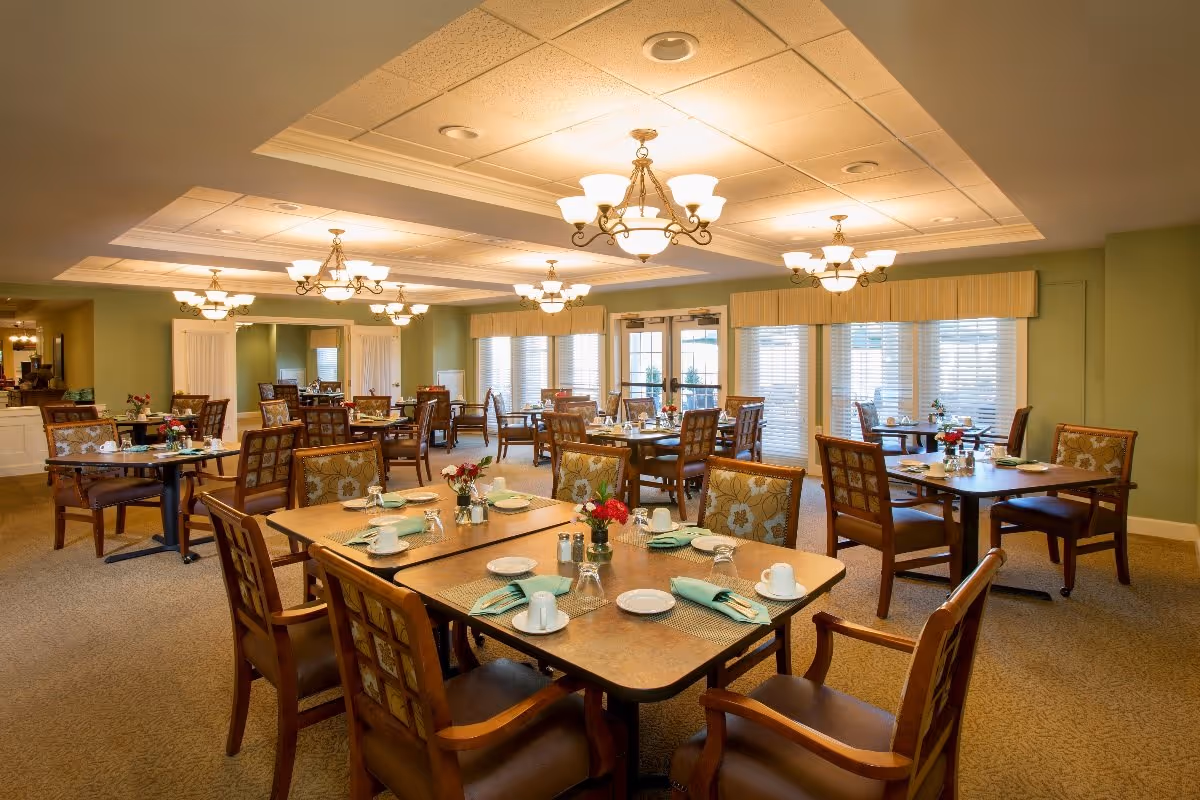 A spacious dining room in Crescent Point at Niantic with multiple wooden tables and chairs arranged neatly. Each table is set with plates, cups, napkins, and silverware. The room is well-lit with elegant chandeliers hanging from the ceiling and large windows with blinds allowing natural light to enter. The walls are painted a soft green color, and the carpet is beige.
