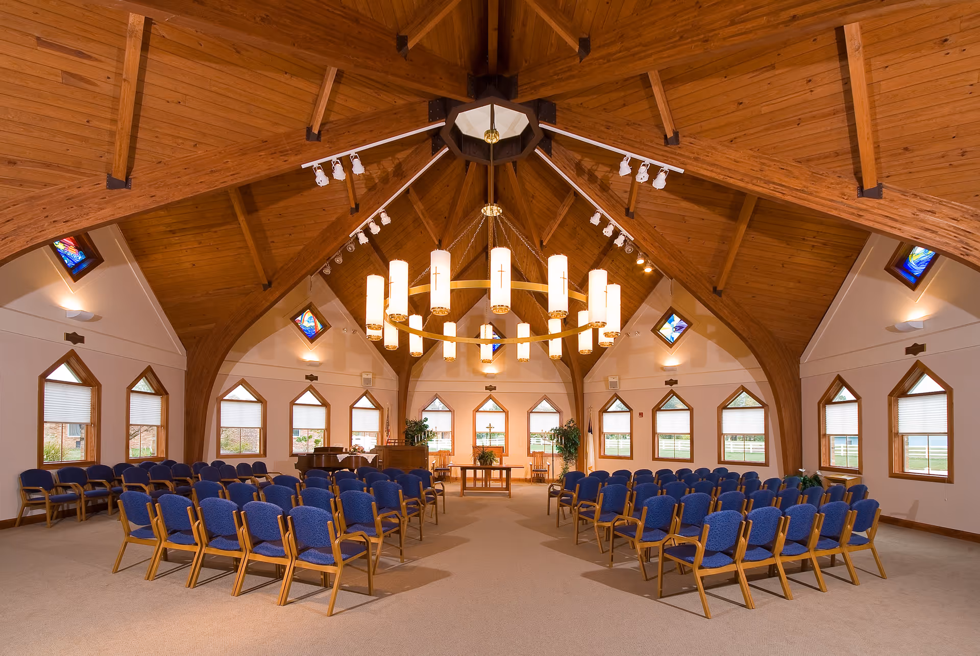 Interior view of a chapel with wooden vaulted ceiling, a large circular chandelier with multiple lights, rows of blue cushioned chairs arranged facing a wooden altar table, and multiple windows with triangular tops and stained glass accents.