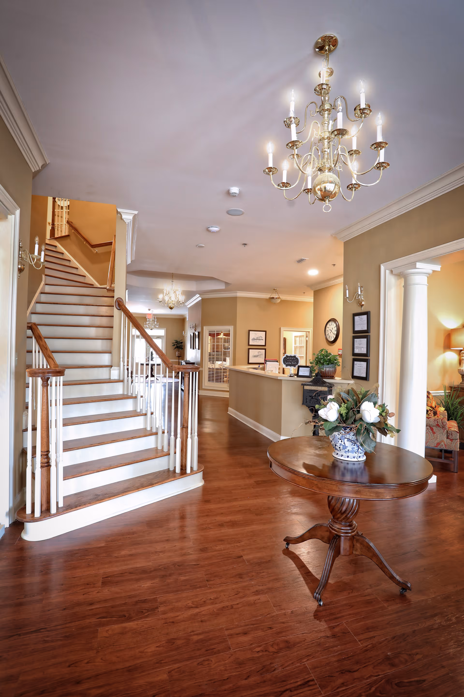 Interior view of a senior care facility featuring a wooden staircase with white railings, a round wooden table with a floral arrangement in a blue and white vase, chandeliers hanging from the ceiling, and a reception desk in the background with framed pictures and a clock on the wall.