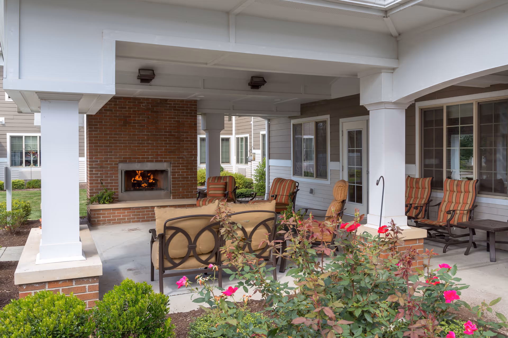 Covered outdoor seating area with cushioned chairs and a sofa arranged around a brick fireplace with a fire burning. The area is surrounded by greenery and flowering plants, with a building featuring windows and doors in the background.
