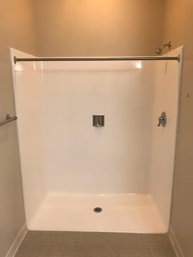 A clean, empty shower stall with beige walls, a silver showerhead, a silver control handle, a soap holder, and a silver towel bar on the left wall.