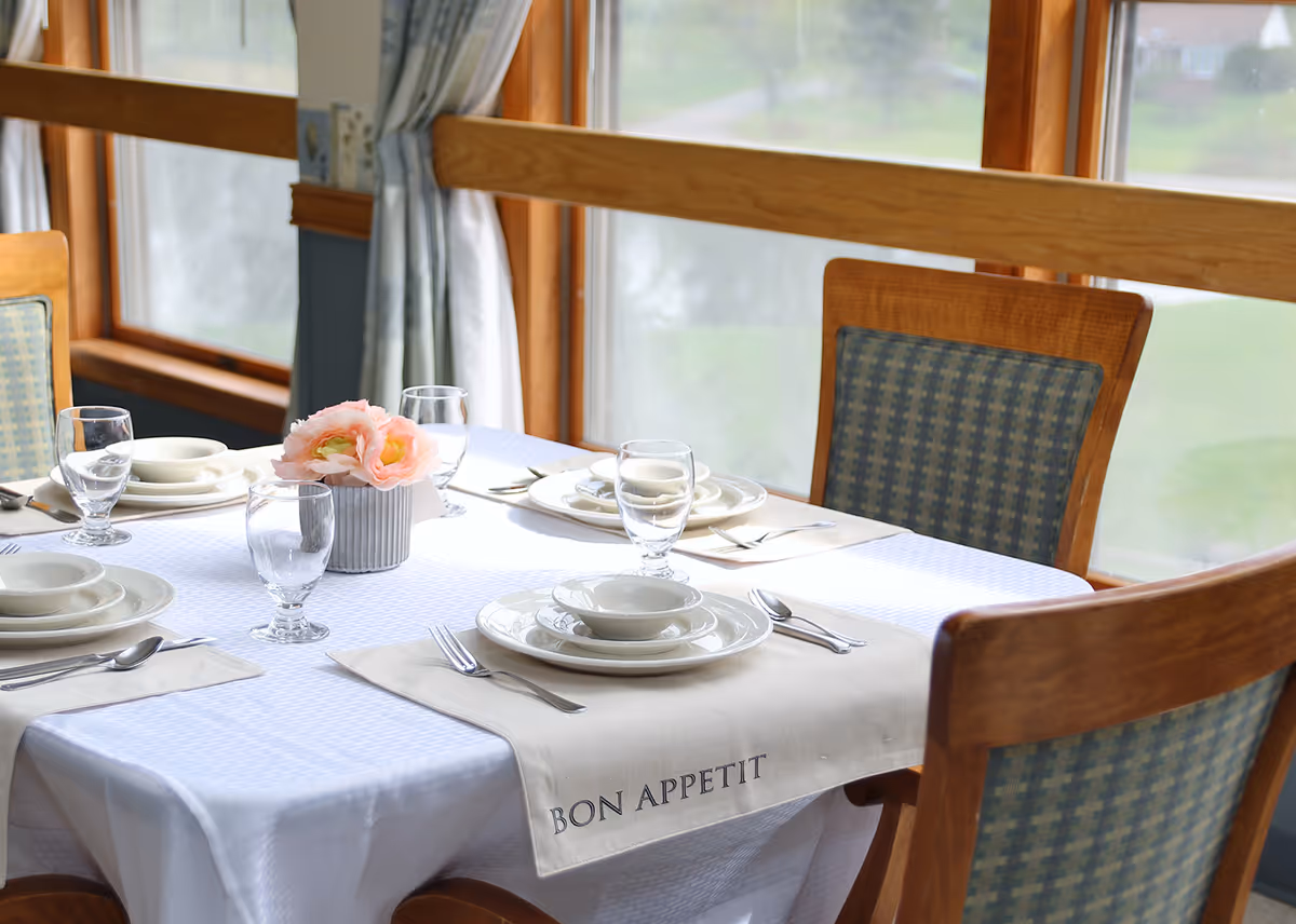 A dining table set with a white tablecloth, place settings, glassware, and a small flower centerpiece by large windows.
