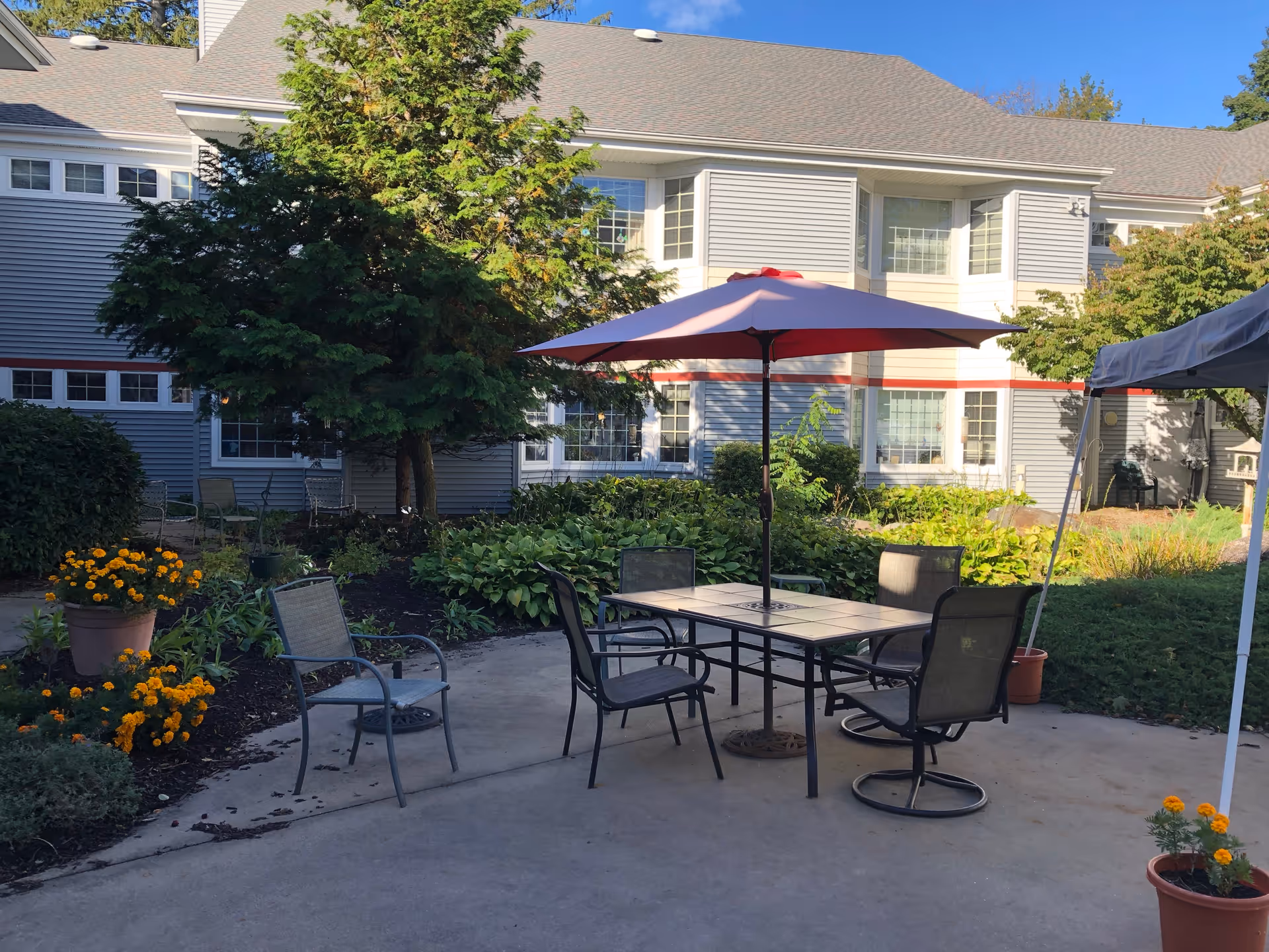 Outdoor patio courtyard with a table, umbrella and chairs in front of a two-story gray building surrounded by plants and potted flowers.