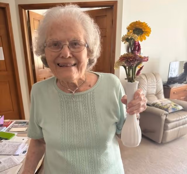 An elderly woman with white hair and glasses smiles while holding a white vase with colorful flowers in a cozy living room. Behind her, there is a beige recliner chair with a floral pillow, a wooden cabinet with a TV, and wooden doors.