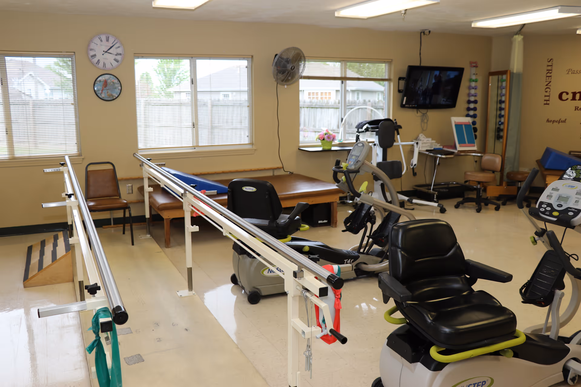 Therapy/exercise room with parallel walking bars, recumbent exercise bikes, and therapy tables beneath windows and wall clocks.