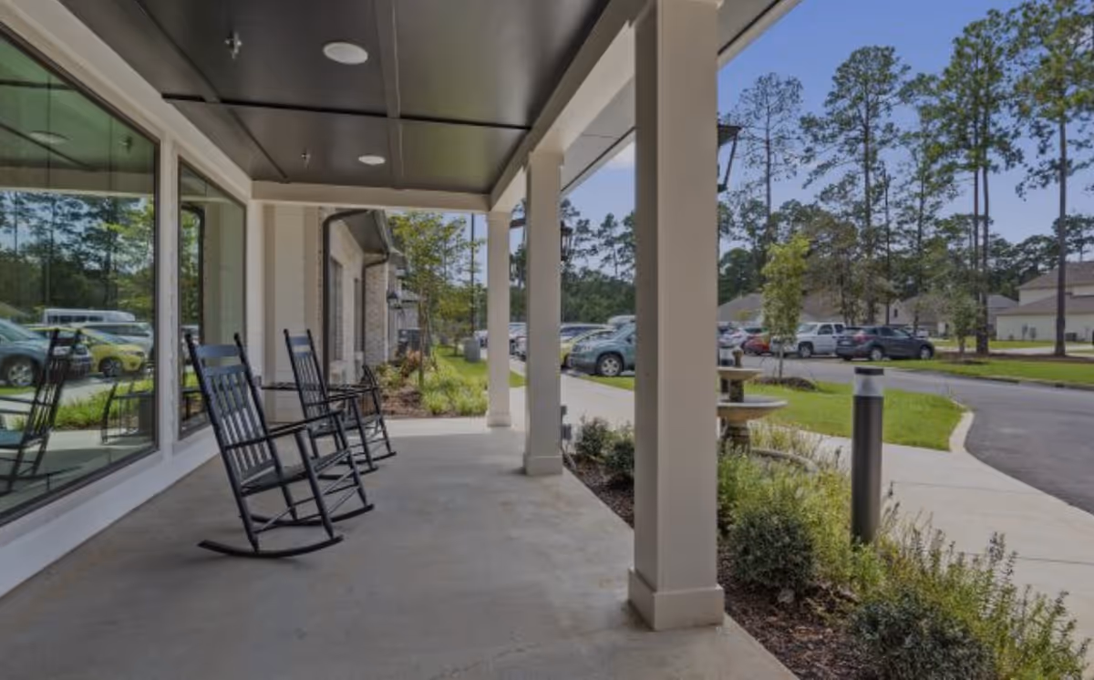 Covered outdoor patio area with black rocking chairs along the side of a building, overlooking a parking lot with cars and surrounded by trees and greenery.