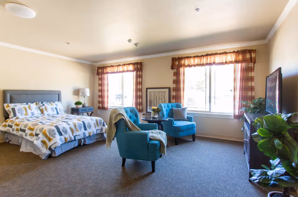 A bright and spacious bedroom in an assisted living facility featuring a bed with patterned bedding, two blue armchairs with a small round table between them, a TV on a stand, and large windows with plaid curtains letting in natural light.