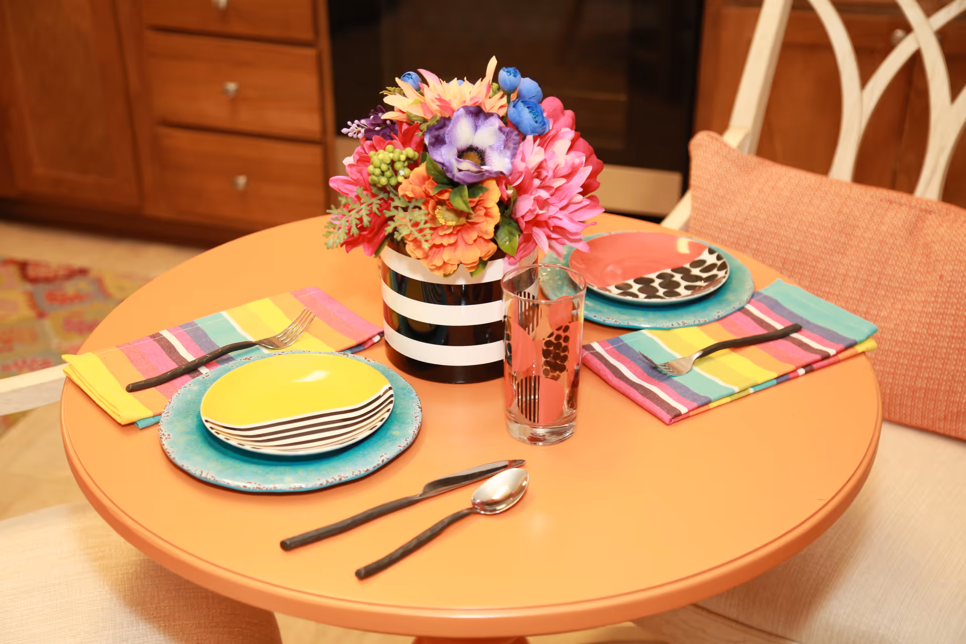 A round orange dining table set for two with colorful striped napkins, mismatched plates in yellow, pink, and black-and-white patterns, and black-handled cutlery. A black and white striped vase with a vibrant bouquet of artificial flowers is placed in the center. The background shows wooden kitchen cabinets and a cushioned chair with an orange pillow.