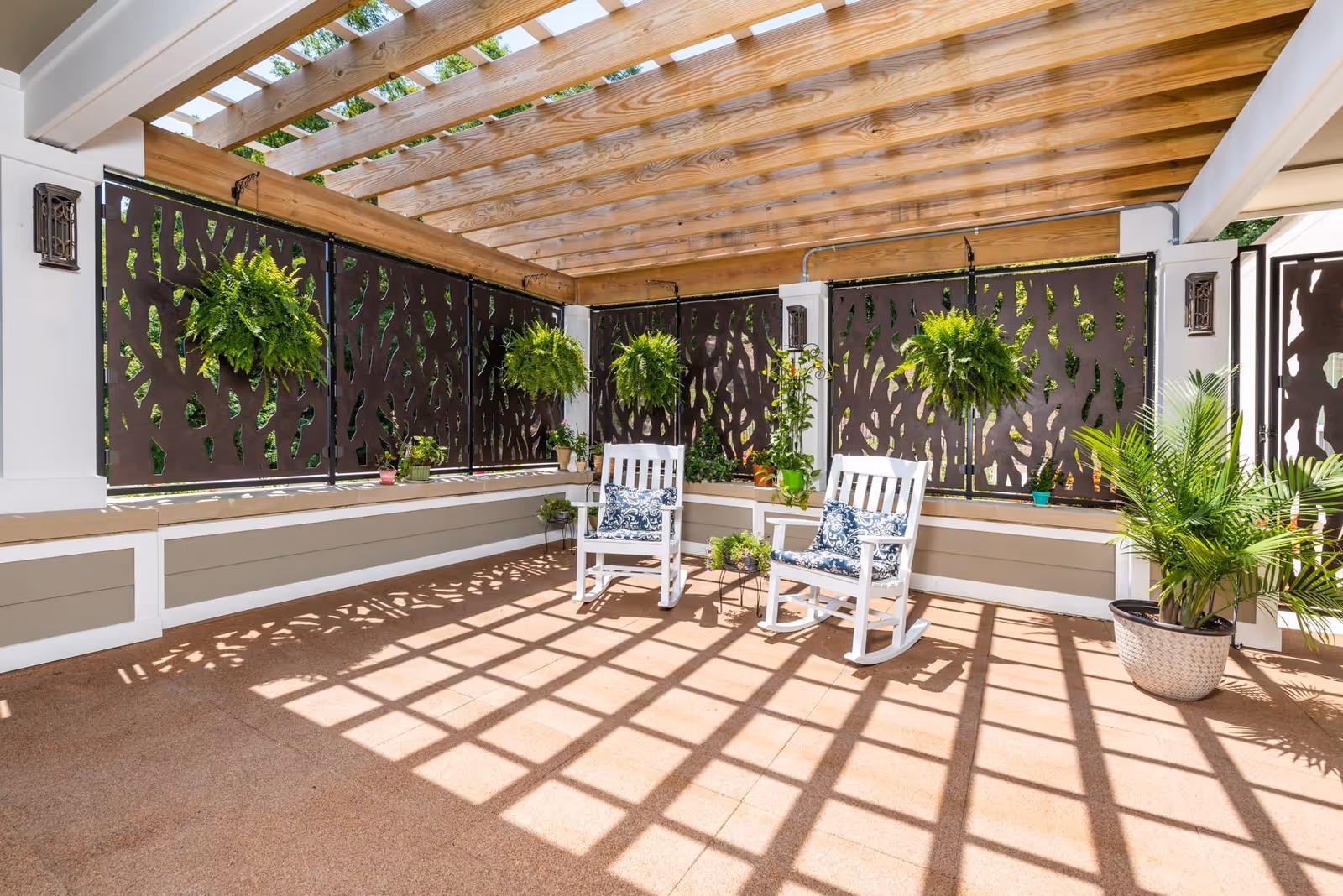 A bright outdoor patio area with a wooden pergola overhead casting shadows on the floor. The space features two white rocking chairs with patterned cushions, several hanging and potted green plants, and decorative privacy panels along the walls.