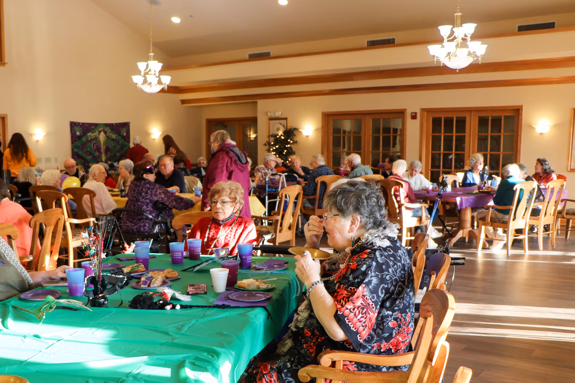 A large dining room filled with elderly people sitting at tables covered with colorful tablecloths, eating and socializing. The room has wooden chairs, chandeliers, and large windows letting in natural light. The atmosphere appears warm and communal.