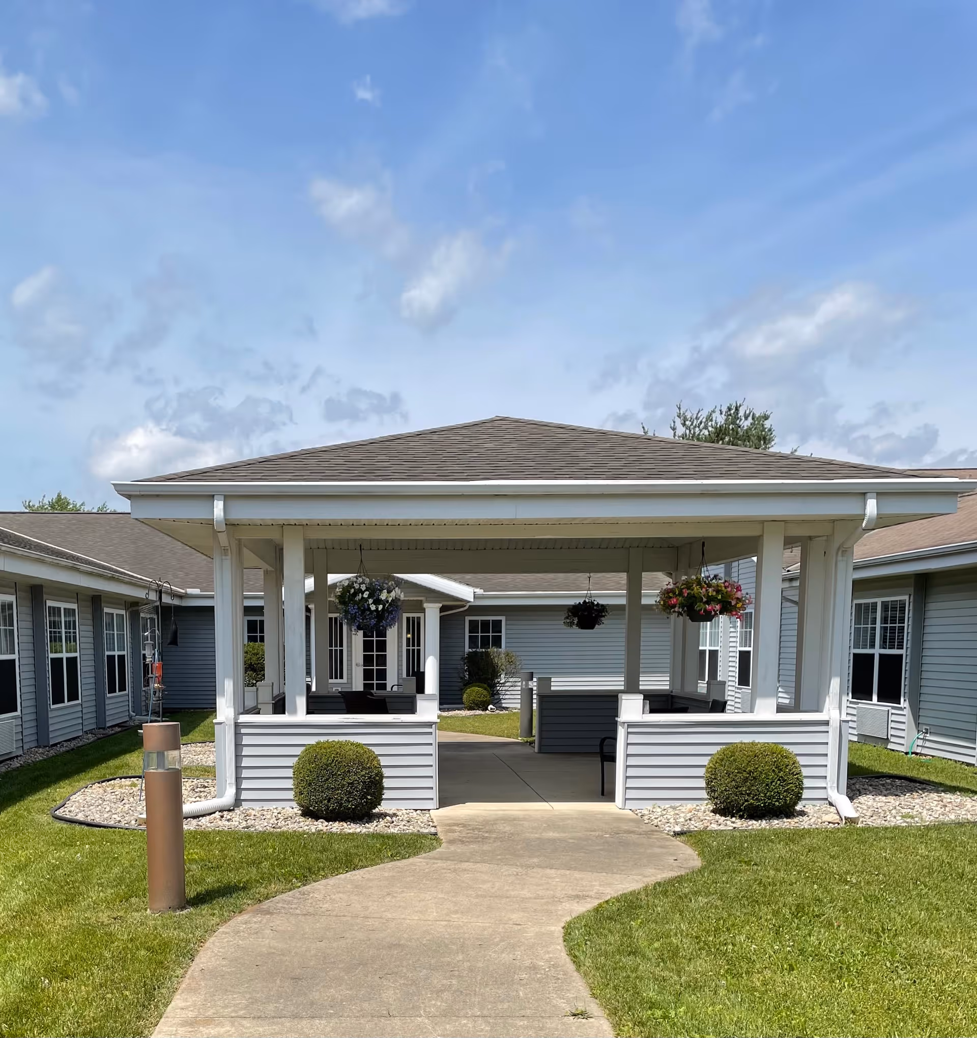 Outdoor covered seating area with hanging flower baskets, surrounded by a well-maintained lawn and bushes, located within a courtyard of a senior living facility with gray siding buildings under a blue sky with some clouds.