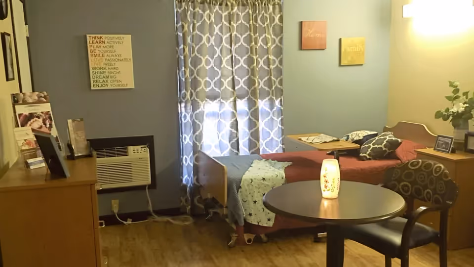 A cozy bedroom in a senior living facility with a single bed covered in red and blue bedding, a small round table with a lit decorative lamp, a patterned chair, a dresser with framed photos and brochures, a window with patterned curtains, and motivational wall art.