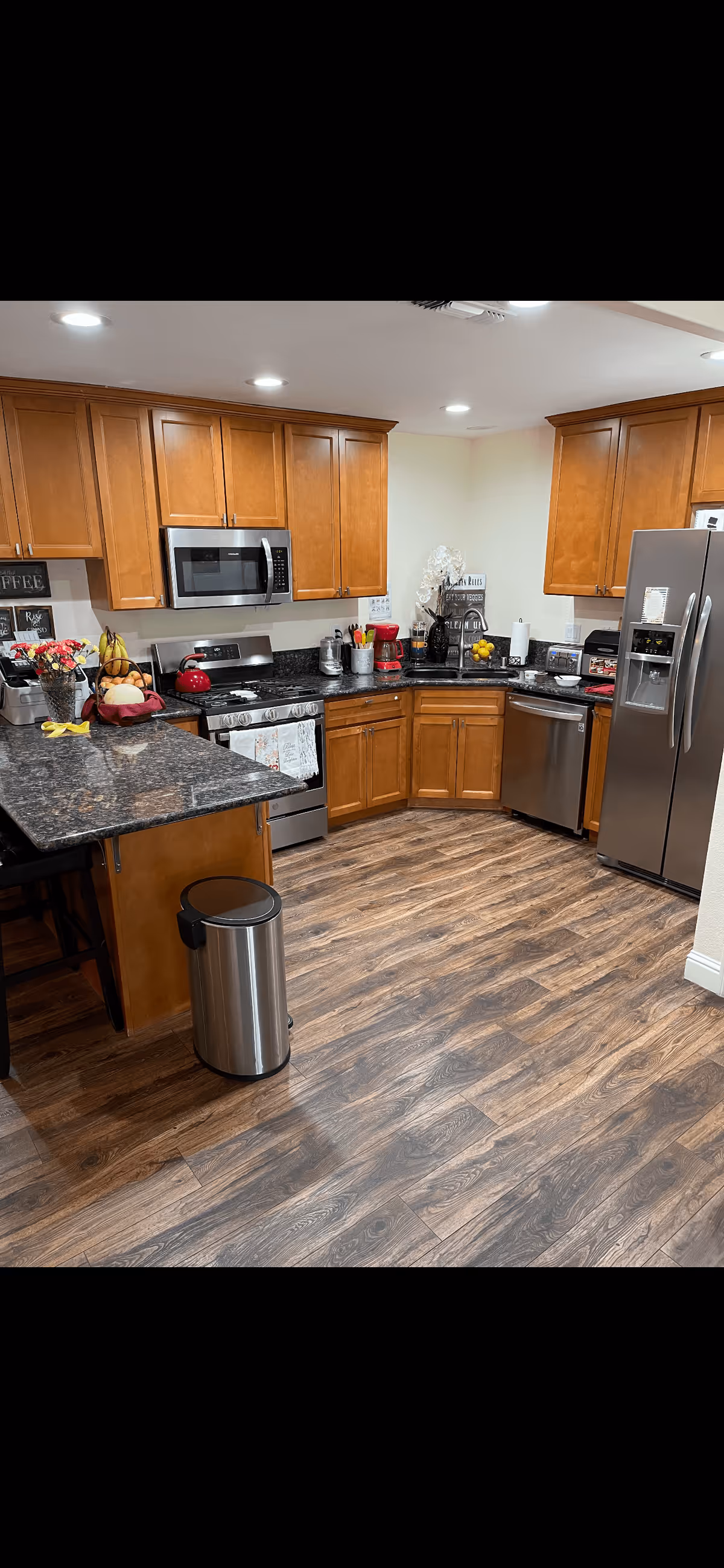 A modern kitchen with wooden cabinets, stainless steel appliances including a refrigerator, microwave, stove, and dishwasher. The kitchen has dark granite countertops, a wooden floor, and various kitchen items such as a red kettle, coffee maker, fruit bowl, and utensils on the counters.