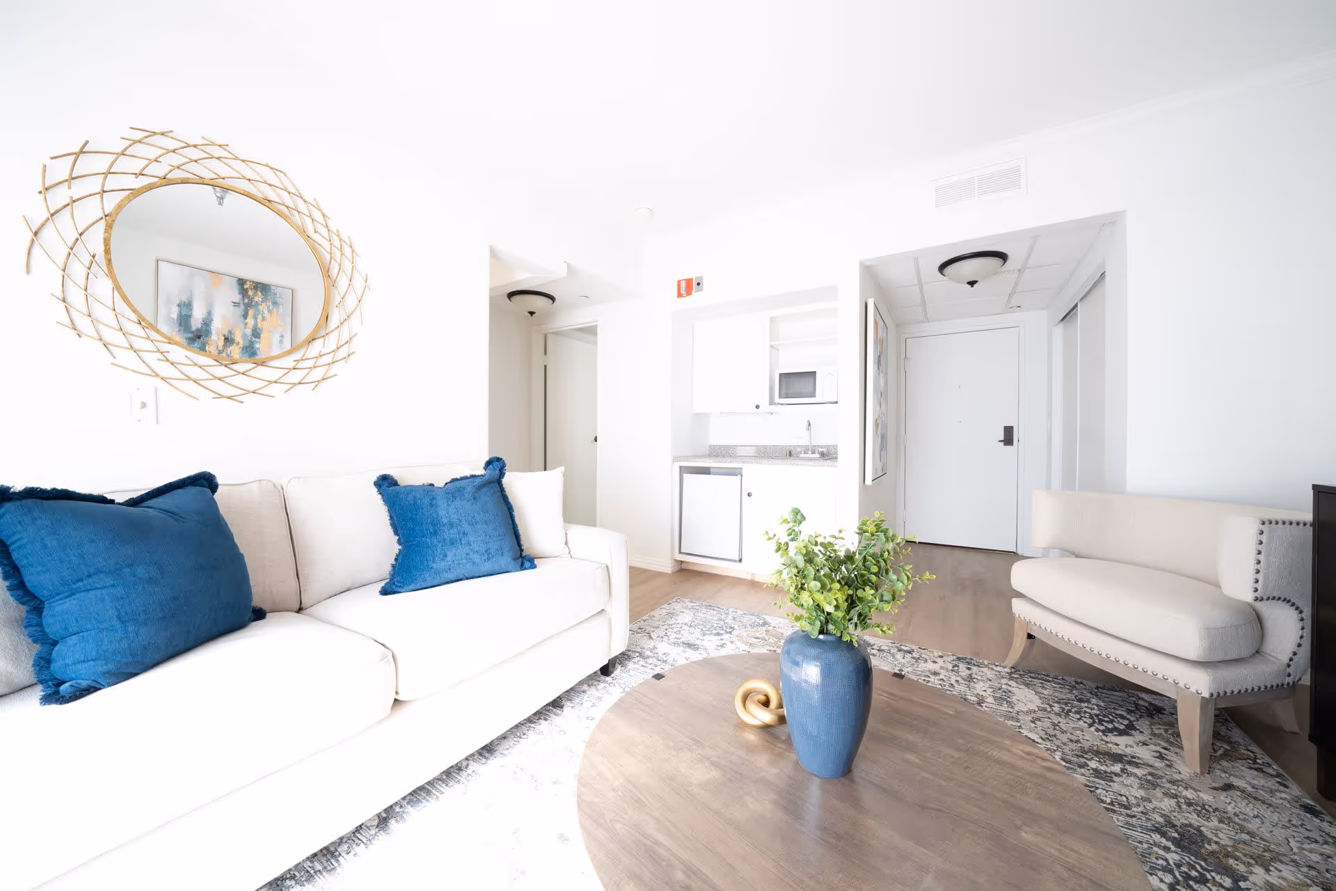 Bright living room with a beige sofa adorned with two blue pillows, a round wooden coffee table with a blue vase and green plant, a beige upholstered bench with nailhead trim, a decorative round mirror with a woven frame on the wall, and a small kitchenette area with a microwave, sink, and mini fridge in the background.