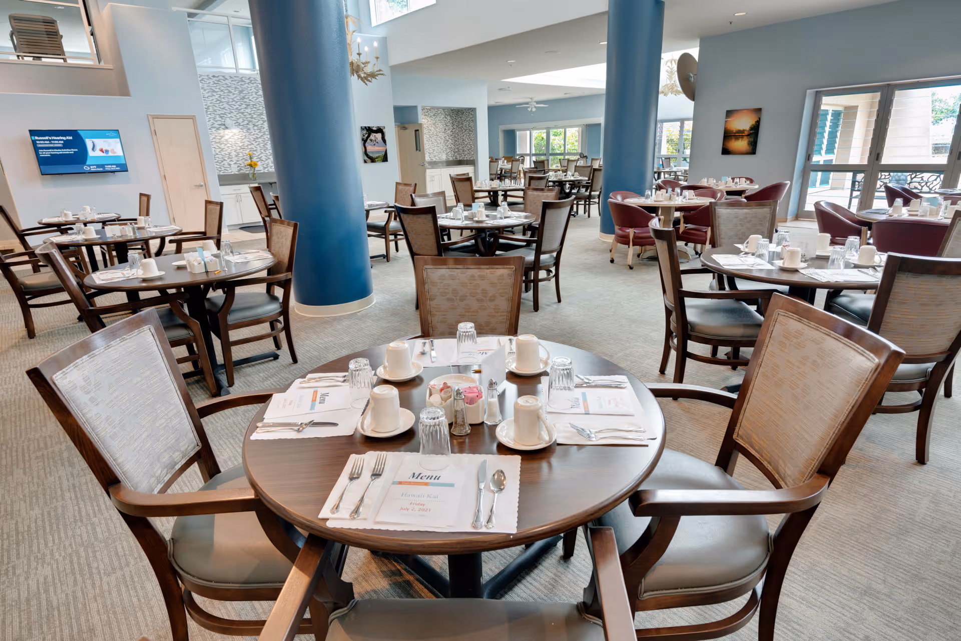 Spacious dining room with round tables set for meals, wooden chairs, place settings, and blue columns.