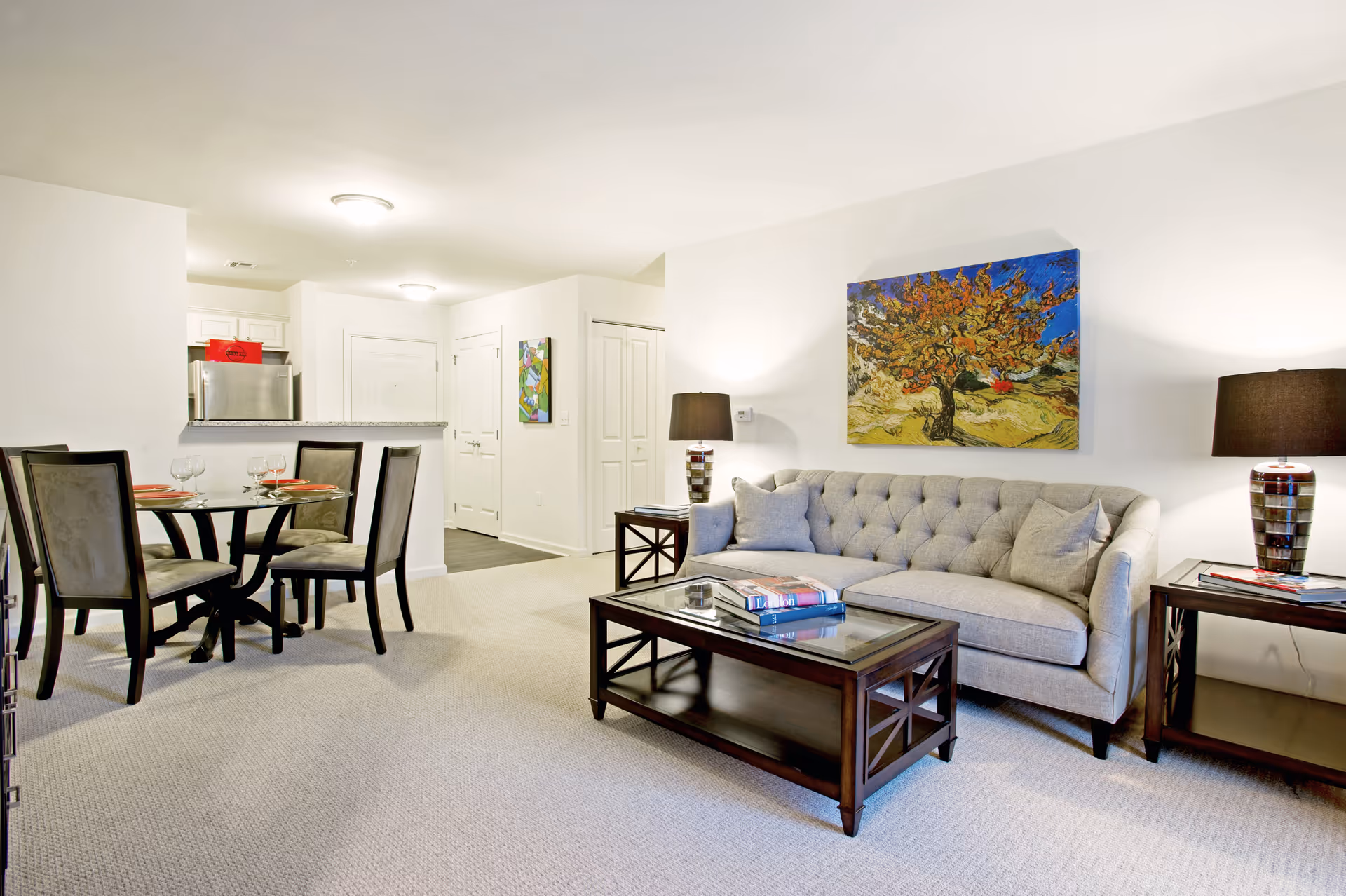 Bright modern living room and dining area with a gray tufted sofa, glass-top coffee table, dining table with four chairs, and colorful wall art.