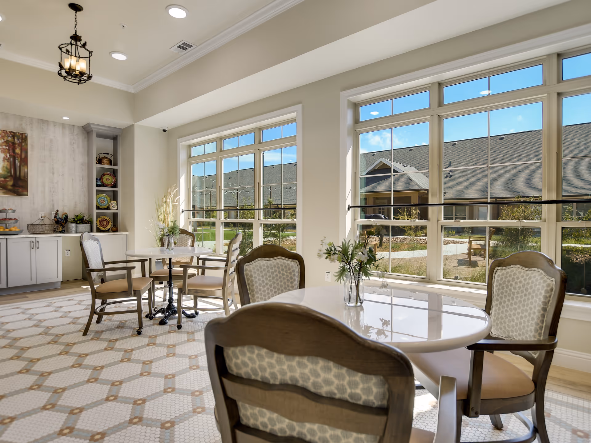 Sunlit dining area with round tables and upholstered chairs beside large windows overlooking a courtyard.