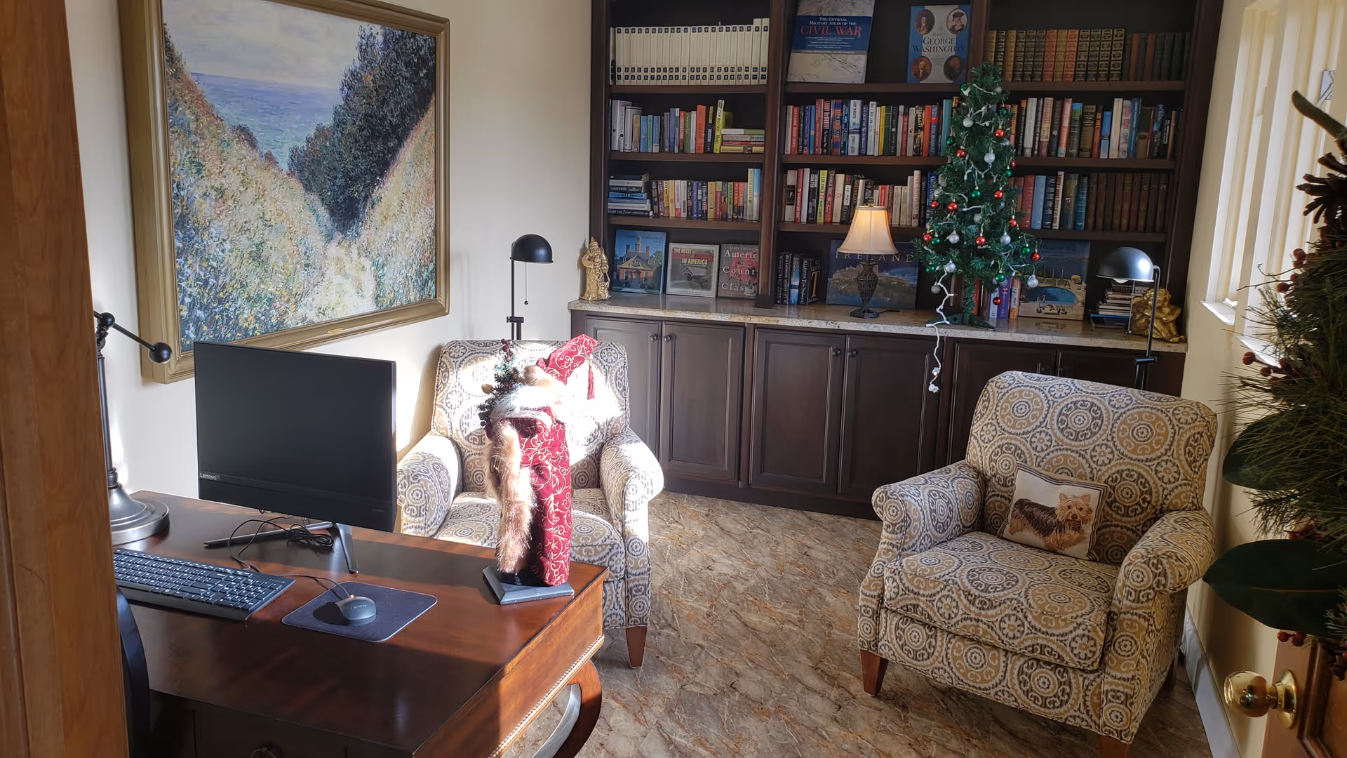 A cozy room with two patterned armchairs, a wooden desk with a computer monitor, keyboard, and mouse. A decorative Santa figure is on the desk. Behind the chairs is a built-in bookshelf filled with books and a small decorated Christmas tree with a lamp. A large framed painting hangs on the wall above one armchair.