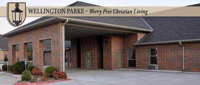 Exterior view of a brick building with a covered entrance and a driveway, featuring landscaping with bushes and a small tree near the entrance. The building has multiple windows and a dark shingled roof.