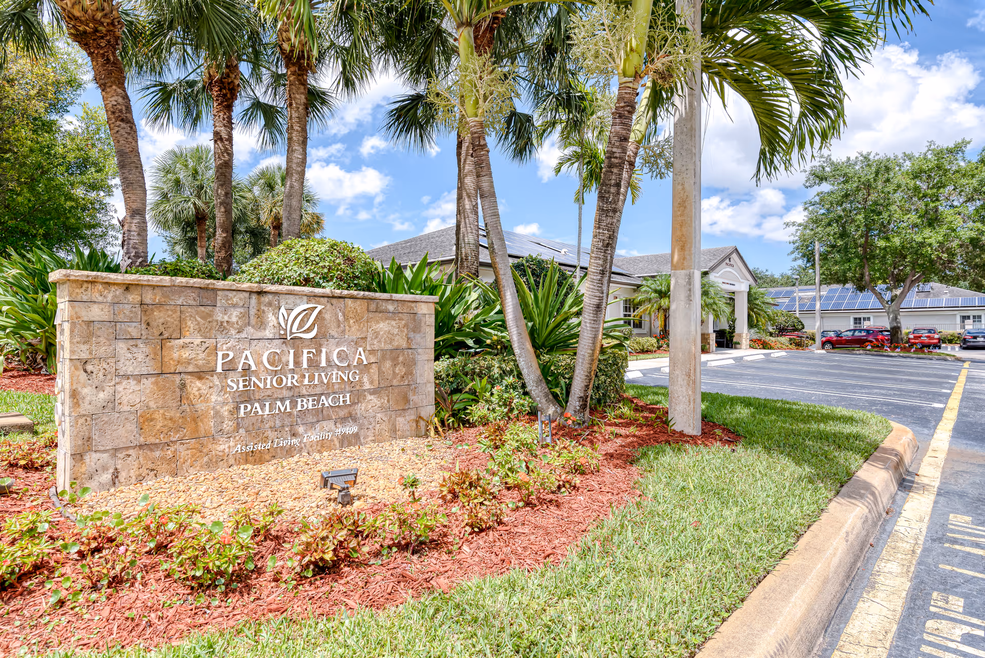 Stone sign for Pacifica Senior Living Palm Beach at a landscaped entrance with palm trees and the facility building and parking lot in the background.