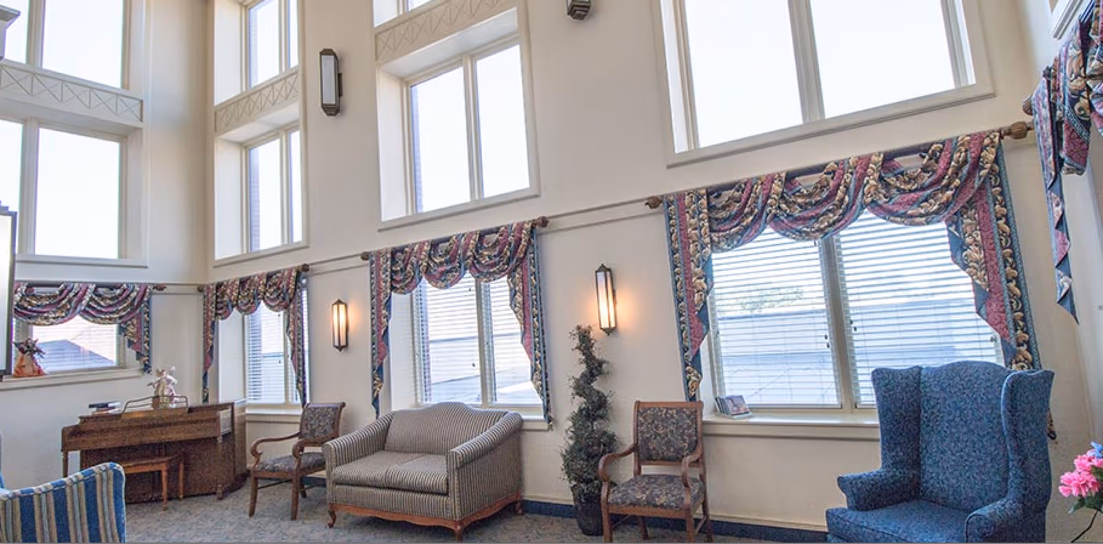 A bright and spacious living room area with large windows covered by patterned valances. The room features a striped loveseat, two wooden chairs with floral upholstery, a blue wingback chair, a small piano with a stool, and a potted plant. The walls are white with decorative wall sconces providing soft lighting.