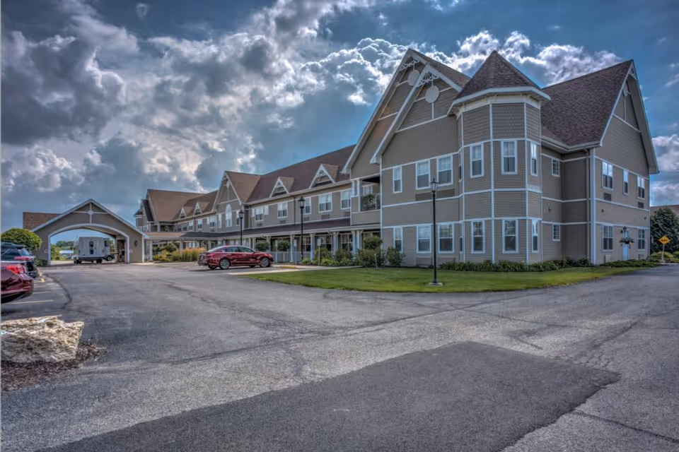 Exterior view of a large, multi-story senior living facility building with beige siding and white trim under a partly cloudy sky. There is a driveway and parking area with several cars parked, and a covered entrance area on the left side of the building.