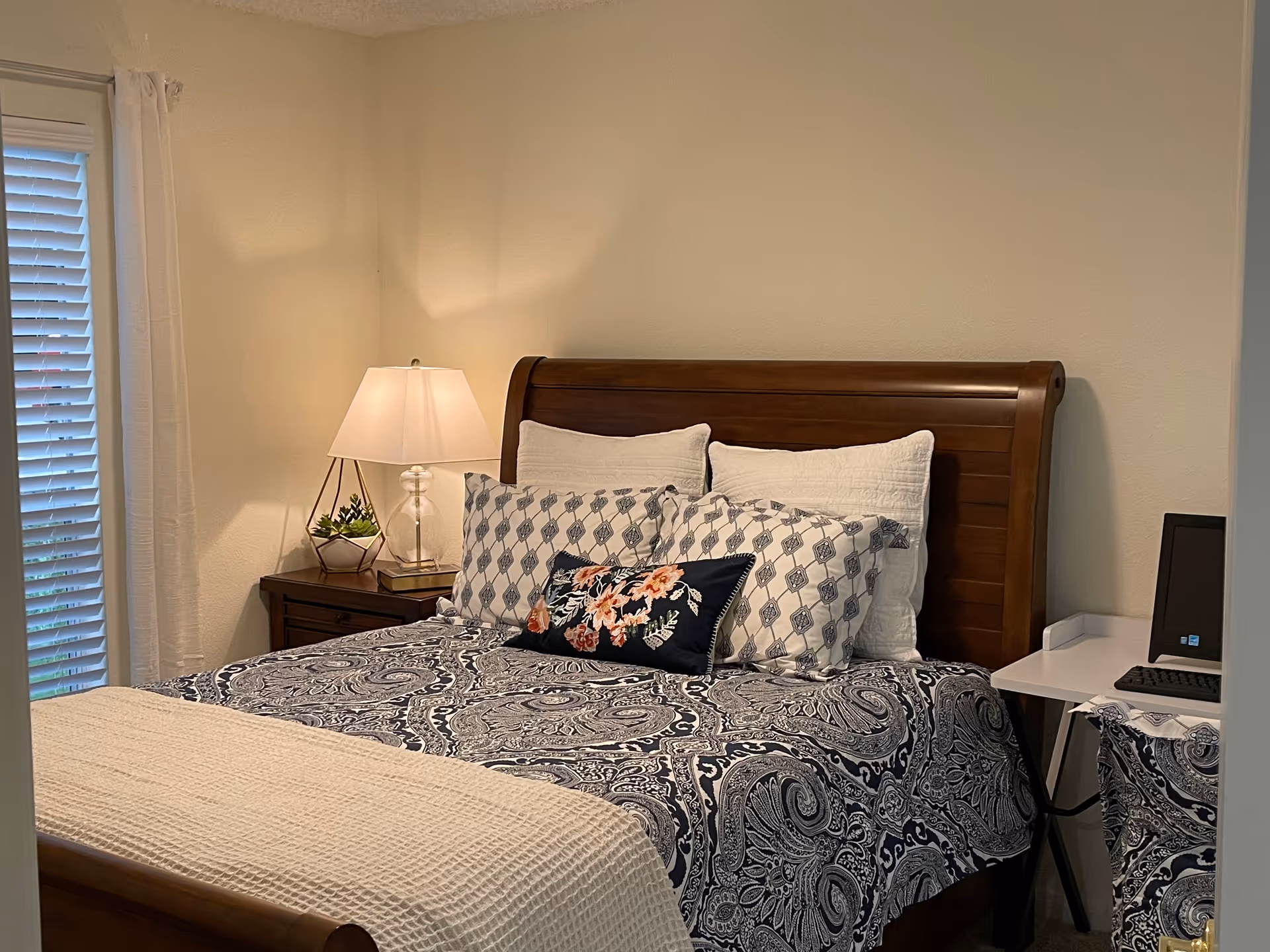 A cozy bedroom with a wooden sleigh bed adorned with patterned blue and white bedding and multiple pillows, a bedside table with a lamp and a small plant, a window with white blinds and curtains, and a small white desk with a computer monitor and keyboard.