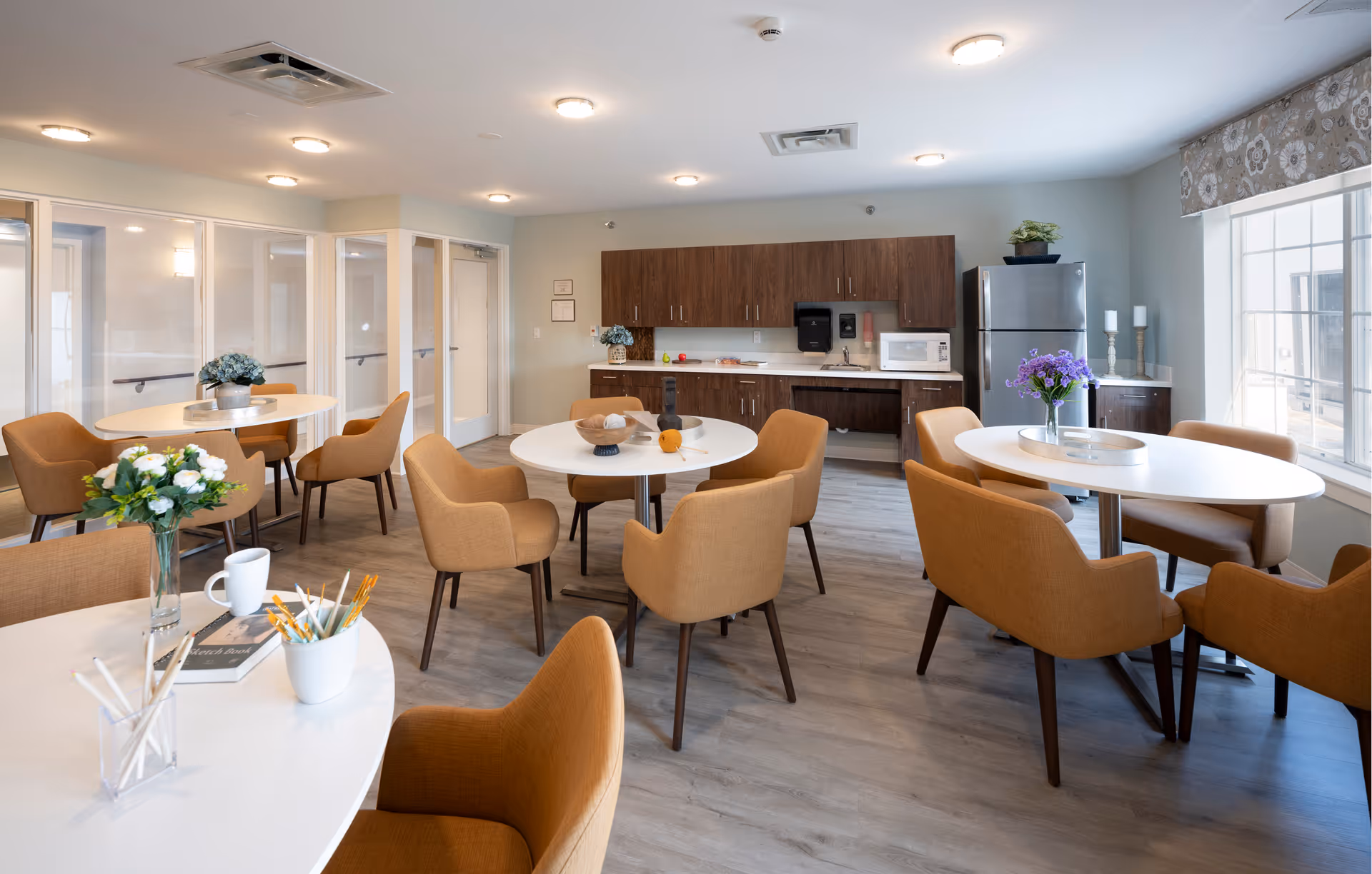 A bright and modern communal dining area in a senior living facility featuring several round white tables surrounded by mustard-colored upholstered chairs. The room has wood flooring, a kitchenette with wooden cabinets, a microwave, and a stainless steel refrigerator. There are decorative flowers on the tables and a large window with a patterned valance letting in natural light.