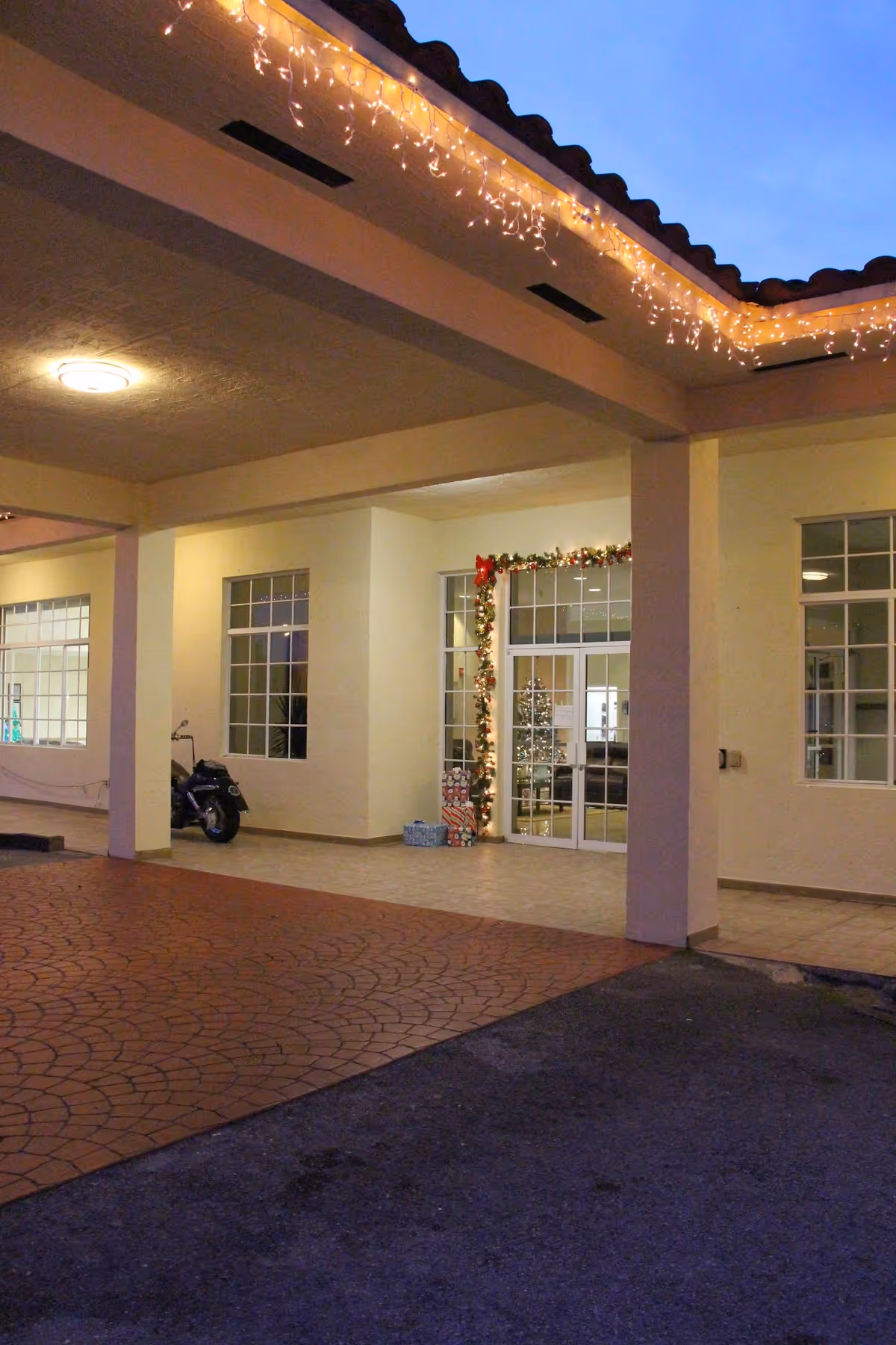 Entrance area of Floridian Gardens Assisted Living Facility at dusk, decorated with Christmas lights and garlands around the door. A Christmas tree and wrapped presents are visible inside through the glass doors. The exterior features a covered walkway with tiled flooring and a parked scooter near a window.