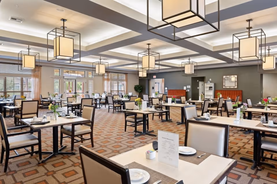 Spacious dining room with multiple tables and chairs arranged neatly. Each table is set with placemats, plates, cups, and small flower vases. The room features large windows with sheer curtains allowing natural light to enter. Modern square pendant lights hang from the ceiling, and there is a patterned carpet on the floor.