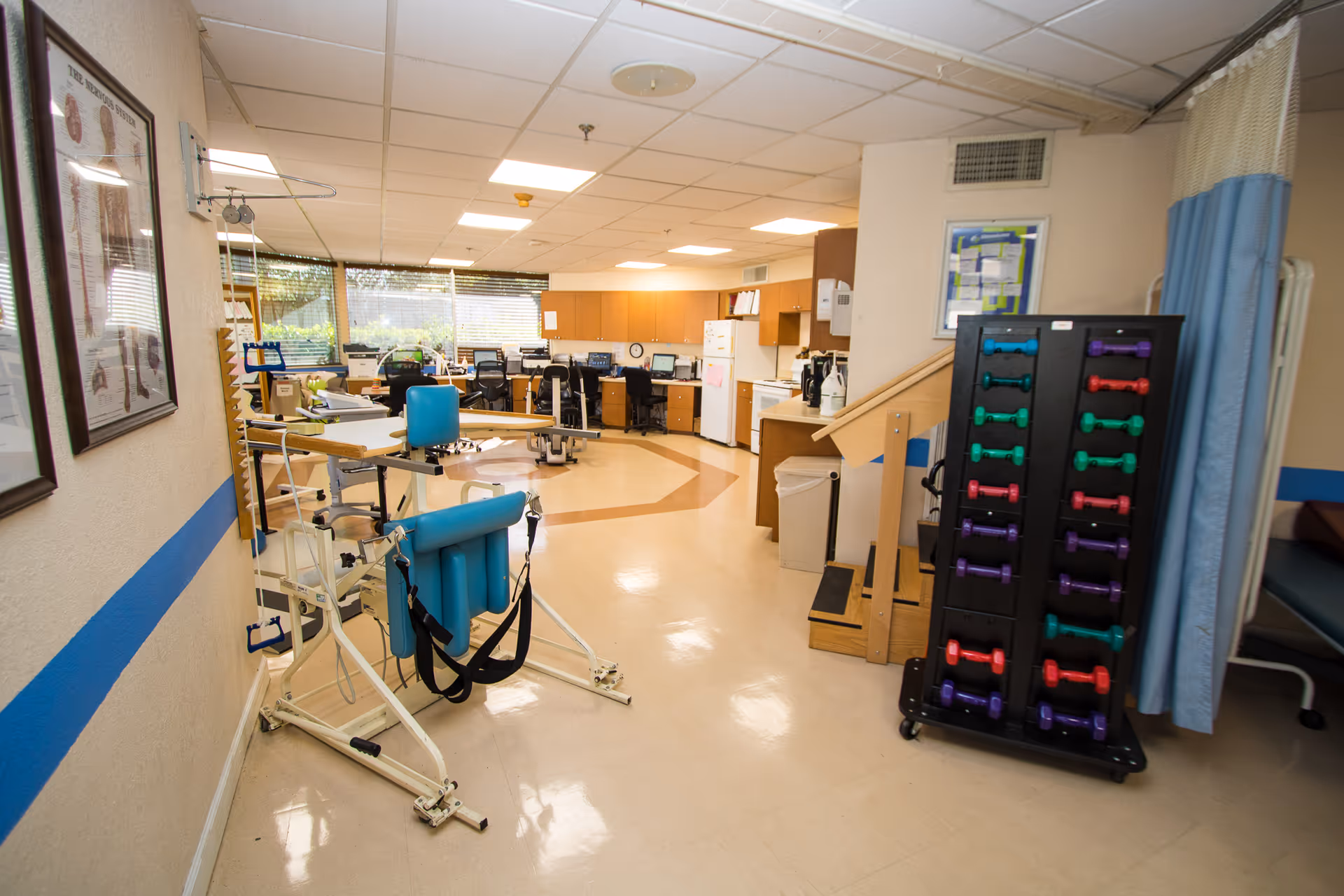 Interior view of a rehabilitation or physical therapy room in a senior care facility. The room contains exercise equipment including a blue padded exercise machine and a rack of colorful dumbbells. There are desks and chairs in the background near windows with blinds, and cabinets along the back wall. The floor is clean and the room is well-lit with ceiling lights.
