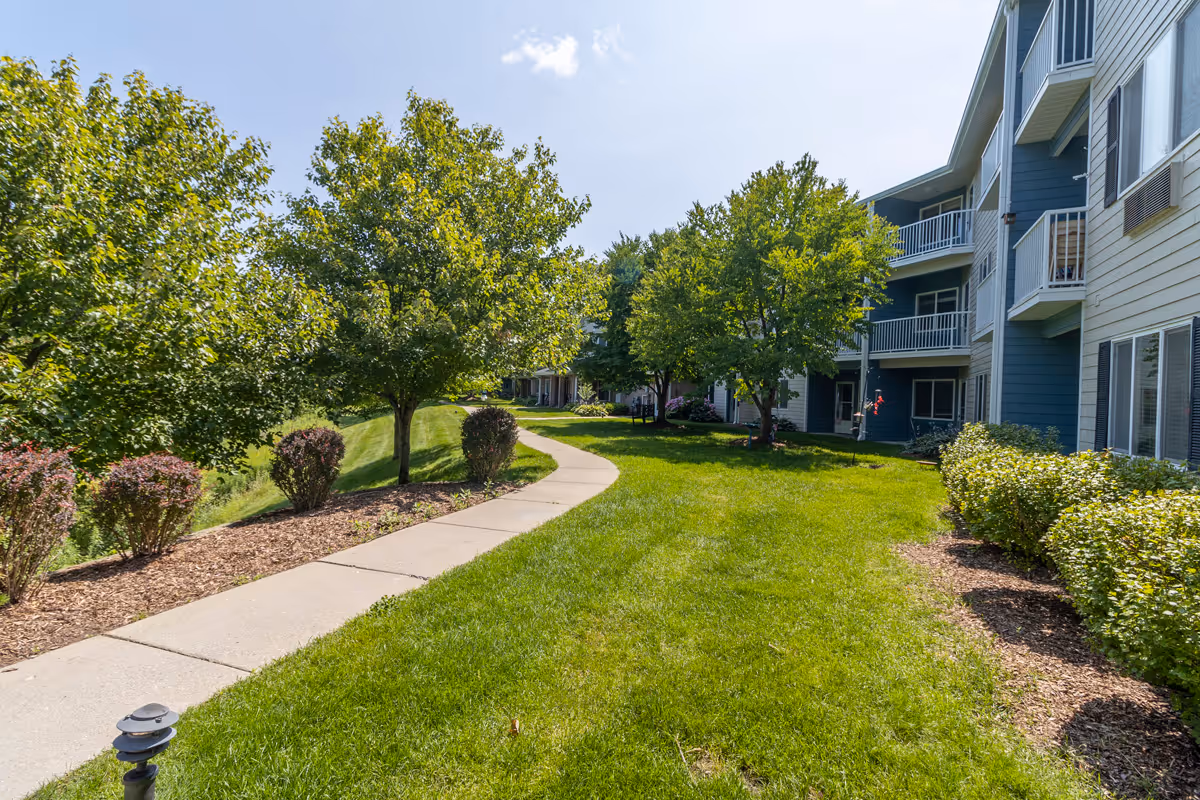 A sunny outdoor pathway winding through a landscaped garden area with green grass, bushes, and trees, adjacent to a multi-story residential building with balconies.