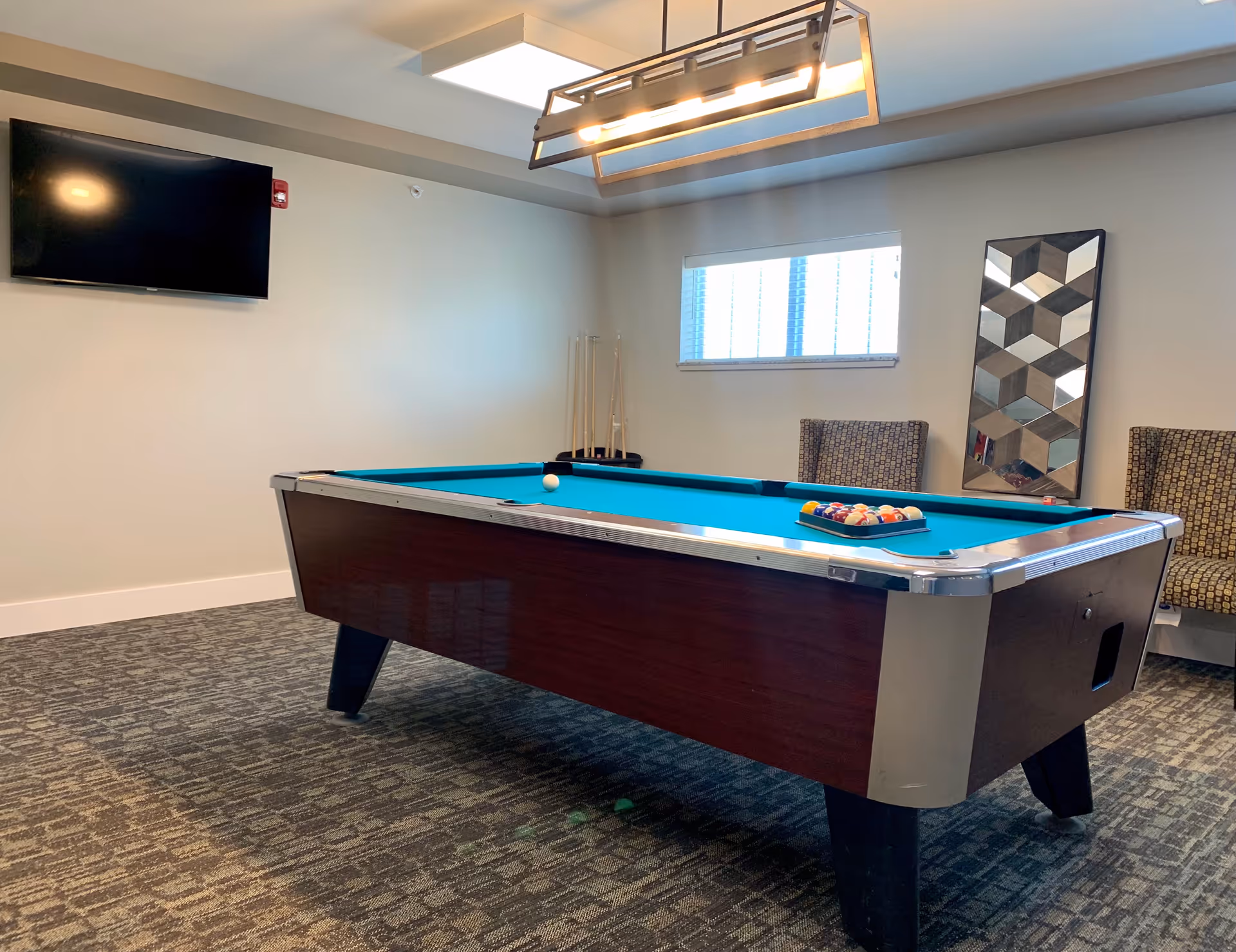 A room with a pool table set up for a game, featuring a rack of billiard balls and a cue ball on the table. The room has patterned carpet, two patterned chairs, a wall-mounted flat screen TV, a window with blinds, a modern rectangular ceiling light fixture, and a decorative mirror on the wall.