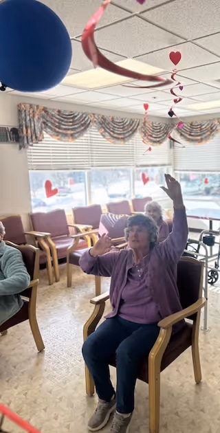 An elderly woman sitting in a chair in a common room decorated with heart-shaped hanging decorations and a blue balloon. She is raising her hands and appears to be engaged in an activity. Other elderly individuals are seated nearby, and the room has large windows with patterned valances letting in natural light.