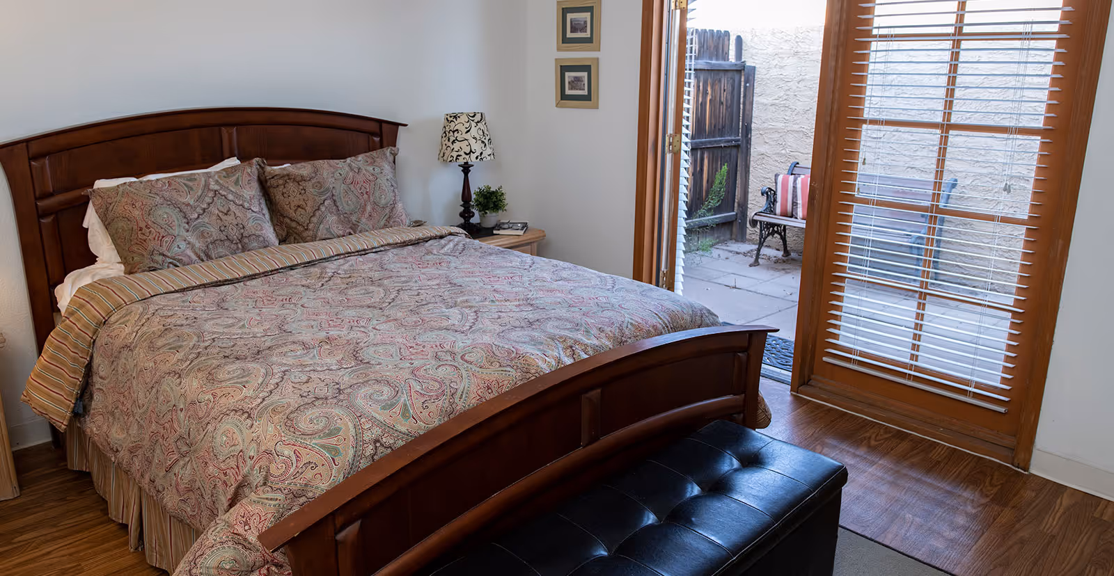 A bedroom with a wooden bed frame and paisley patterned bedding. There is a nightstand with a lamp and a small plant next to the bed. The room has wooden flooring and a door with glass panels and blinds leading to a small outdoor patio with a bench and cushions.