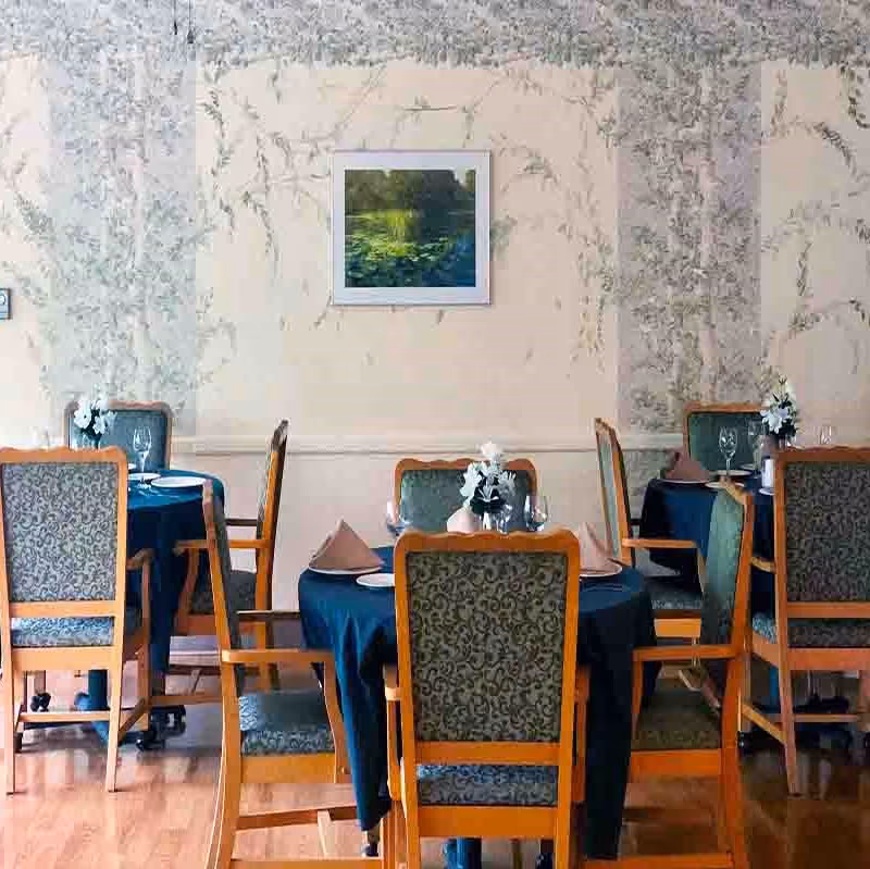 Dining room with round tables covered in dark blue tablecloths, each set with plates, napkins, wine glasses, and small floral centerpieces. Wooden chairs with patterned upholstery surround the tables. The walls have a light-colored floral wallpaper and a framed picture of a pond scene hangs on the wall.