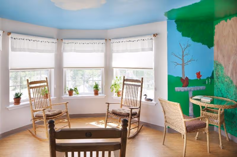 A bright room with three wooden rocking chairs arranged in a semi-circle near a bay window with white blinds. Several small potted plants are placed on the window sill. The walls are painted with a mural depicting a tree, birds, and a birdbath. There is also a small wicker table with two wicker chairs on the right side of the room.